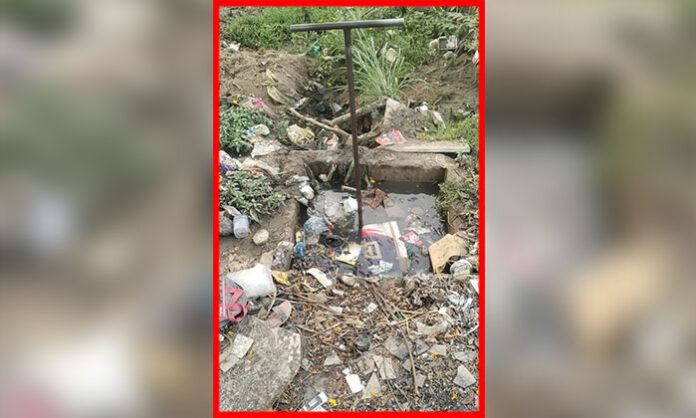 A dirty and clogged manhole filled with stagnant sewage water and garbage near an urban road causing mosquito breeding and health hazards.