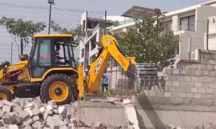 Officials demolishing illegal compound wall structures near Kokapet Lake as part of Hydra-led lake restoration and encroachment removal drive in Hyderabad.